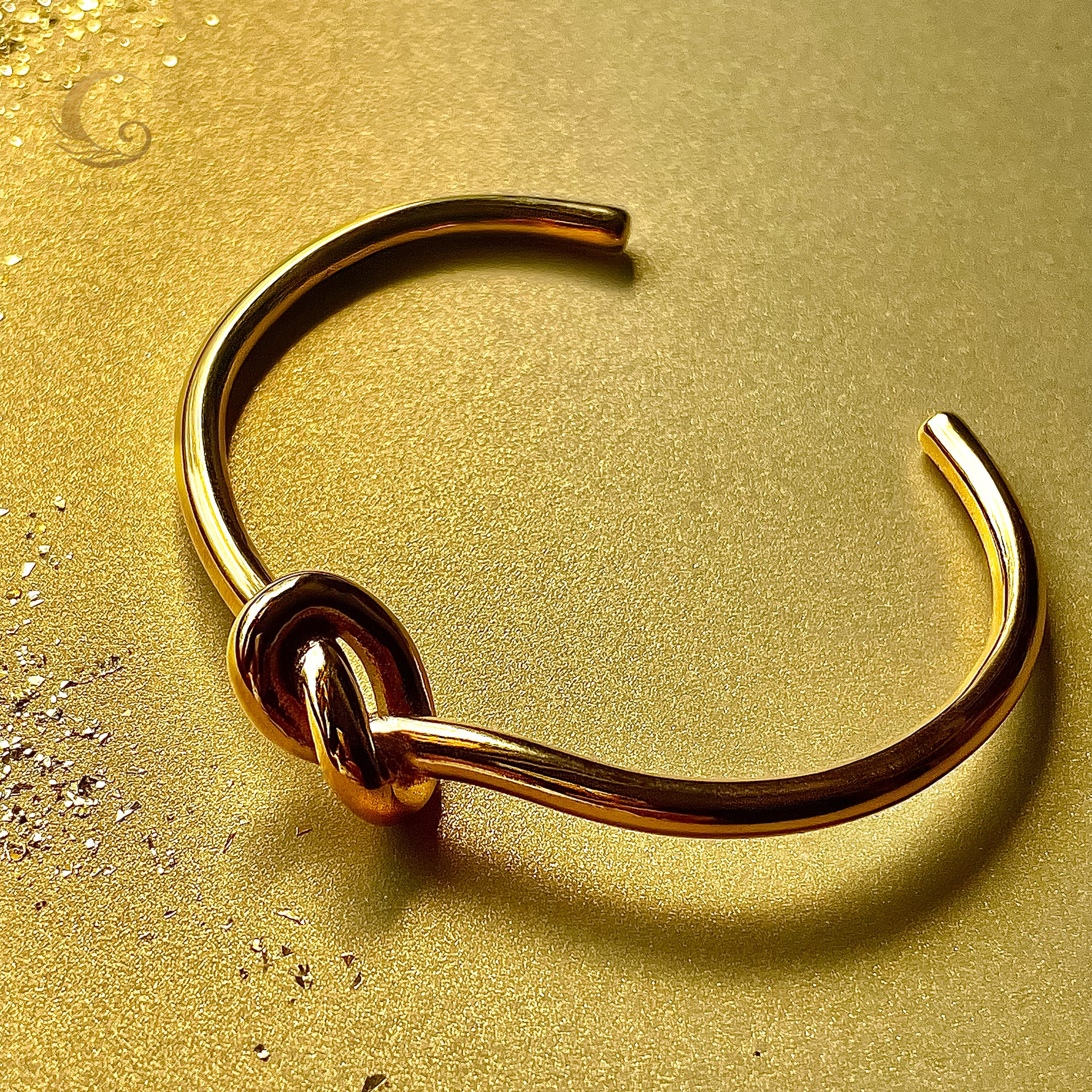 Gold open bracelet with symbolic knot, top view on soft surface, Earth Collection – Avyrona