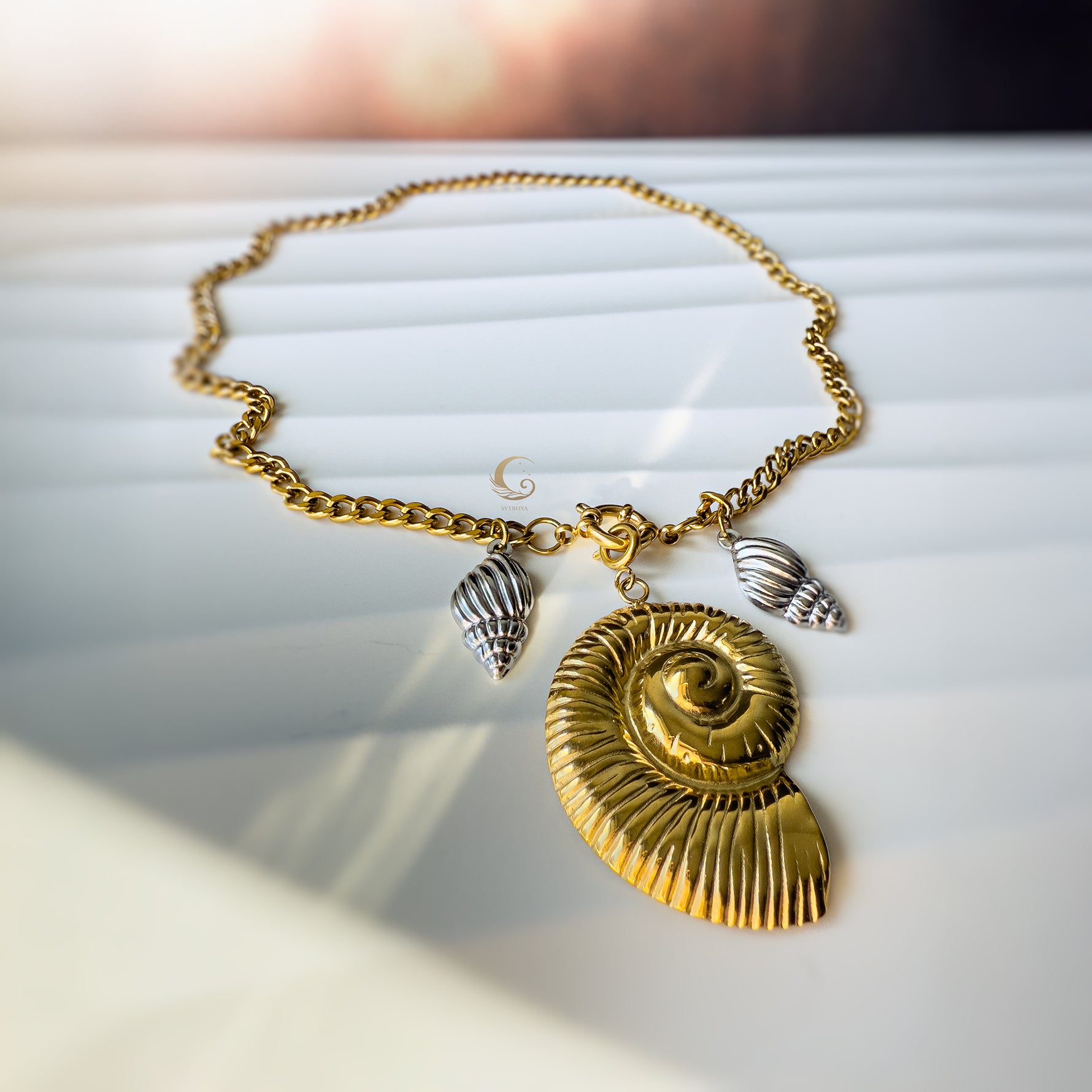 Close-up of the Thalassa necklace with a large gold shell pendant and two silver shell charms, resting on a sunlit white surface.
