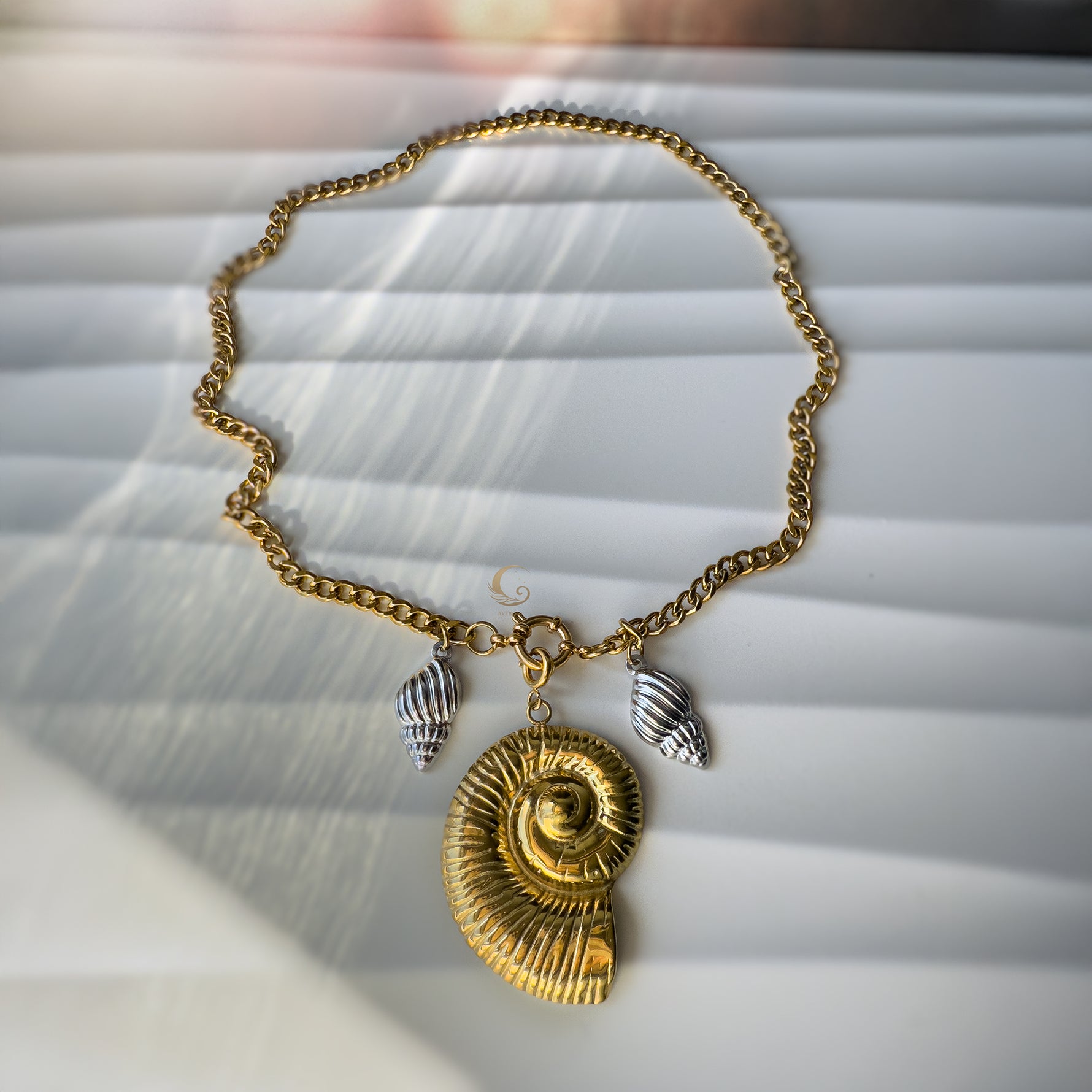 Full layout of the Thalassa necklace with large gold shell and two silver charms, artistically arranged on a striped background.