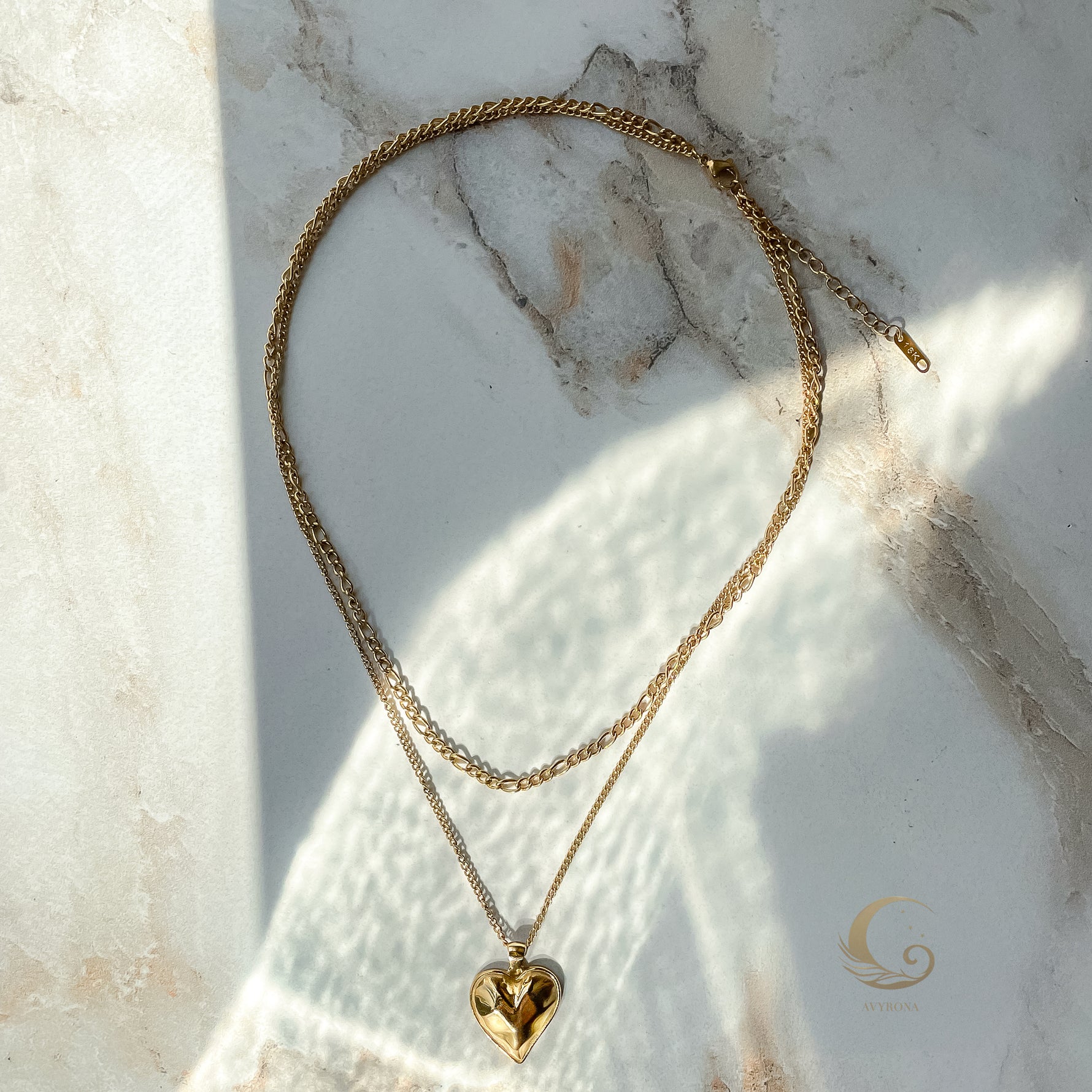 Full view of Vesta gold layered necklace arranged in oval shape on marble surface, styled with soft glow and Avyrona watermark.