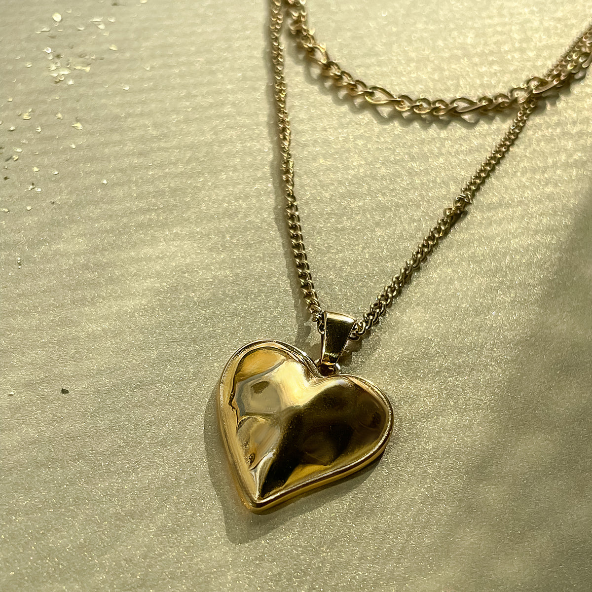 Reflective heart-shaped pendant with double chain displayed in warm light on textured surface, styled in layered formation.