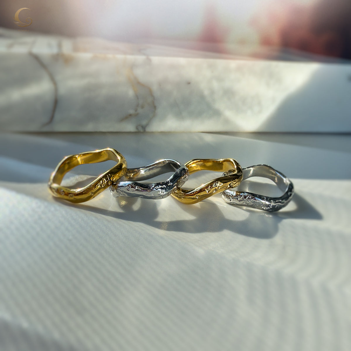 Wavy gold and silver rings resting on white and marble textures, showcasing their minimalist curves.