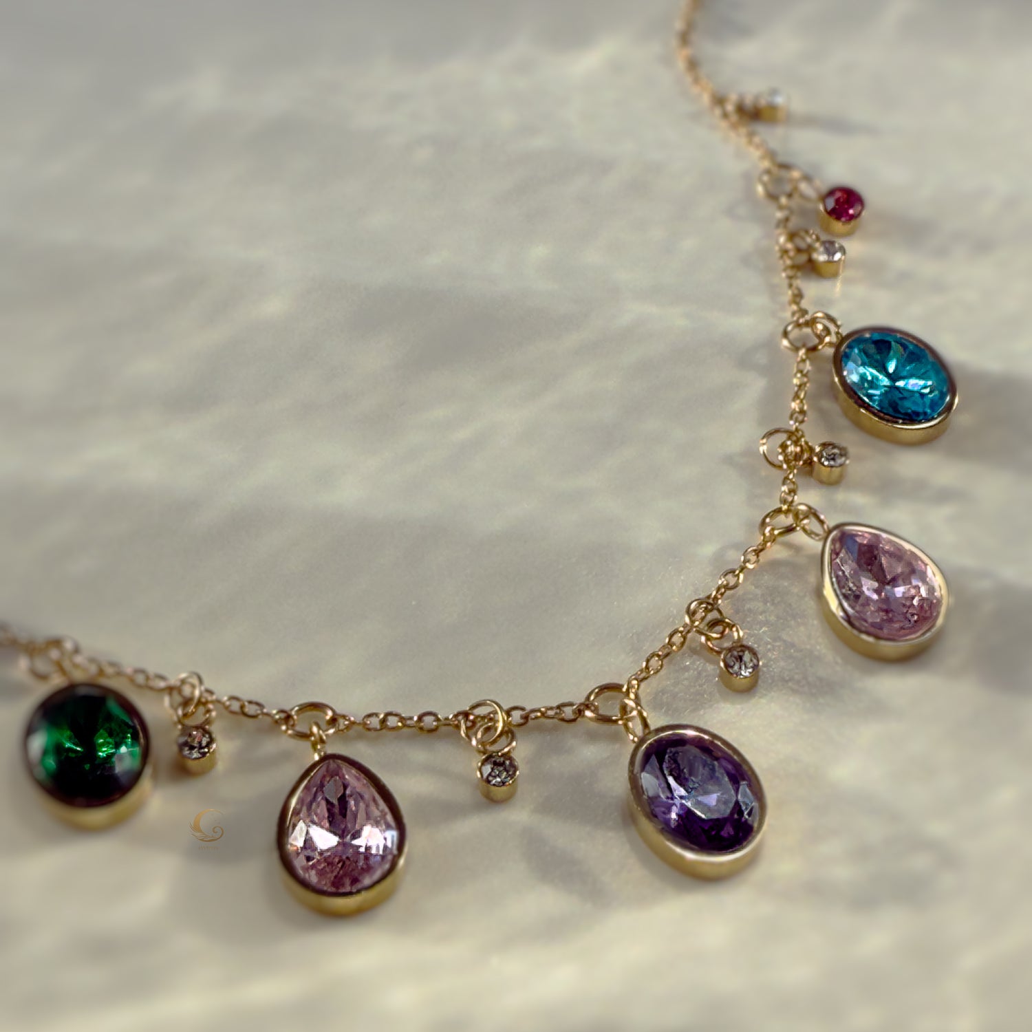 Gold charm necklace with colorful teardrop and round crystals displayed on a smooth white stone surface