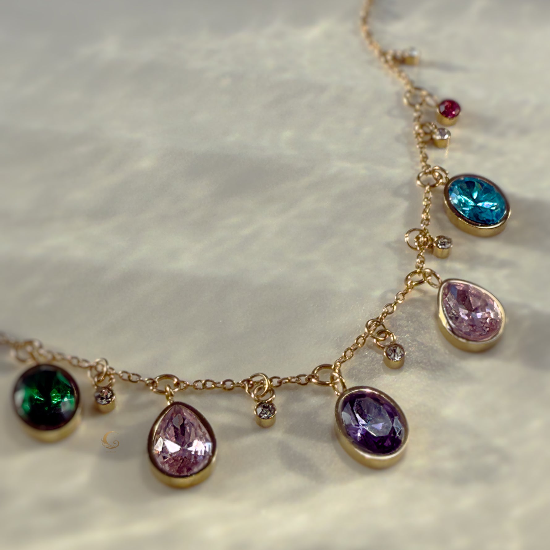 Gold charm necklace with colorful teardrop and round crystals displayed on a smooth white stone surface